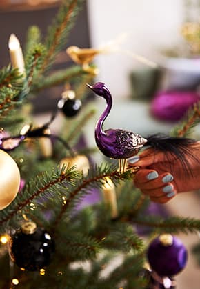 Hand mit hellblau lackierten Nägeln hängt lila Vogel-Glasornament an Weihnachtsbaum mit verschiedenen Kugeln und Lichtern