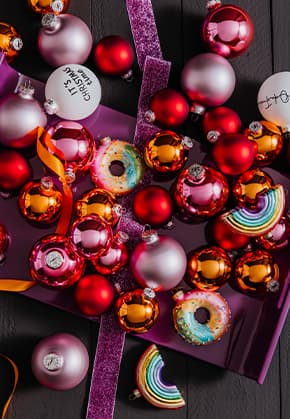 Weihnachtskugeln in rot, orange und silber auf lila Geschenkpapier, bunte Donut- und Regenbogenanhänger dazwischen