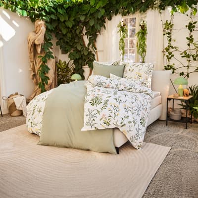 Bett mit geblümter Bettwäsche und grüner Decke in hellem Raum mit vielen Pflanzen und Statue im Hintergrund