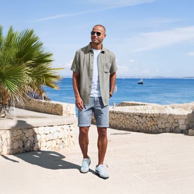 Mann in offenem Hemd und Shorts spaziert an einer Steinmauer mit Meerblick und Palme im Hintergrund