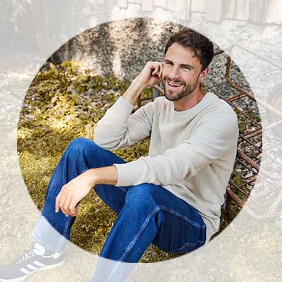 Mann in hellem Pullover und Jeans sitzt lächelnd im Garten auf dem Boden