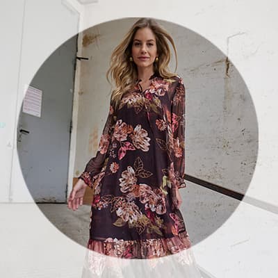 Frau mit langen Haaren in knielangem, dunkel gemustertem Blumenkleid steht in einem hellen Flur und blickt in die Kamera