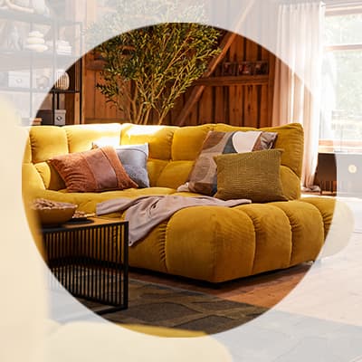 Gelbes Ecksofa mit vielen Kissen und Decke in gemütlichem Wohnzimmer mit Holzregal, Pflanze und großem Fenster