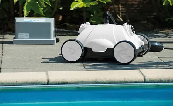 Weißer Poolroboter mit vier Rädern auf grauen Fliesen neben einem Schwimmbecken und einer Steuerbox im Hintergrund