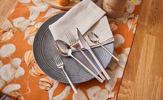 Besteckset auf grauem Teller, umgeben von orangefarbenem Blumenmuster, neben beigem Stoffserviette