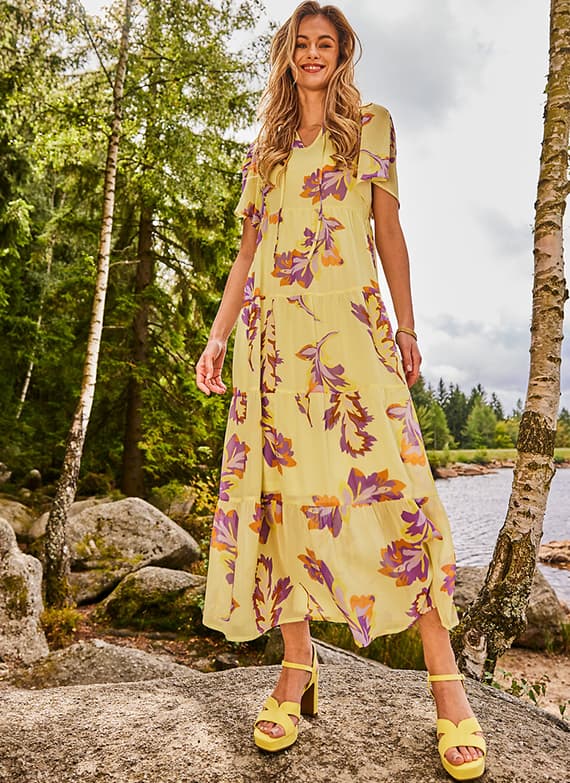 Frau im gelben Maxikleid mit großem Blumenmuster steht auf Felsen am See zwischen Bäumen