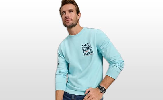 Mann in hellblauem Sweatshirt mit kleinem Schriftprint auf der Brust, eine Hand in der Hosentasche, neutrale Pose