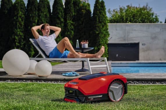 Roter Mähroboter fährt über Rasen, im Hintergrund entspannt ein Mann auf einer Liege am Pool
